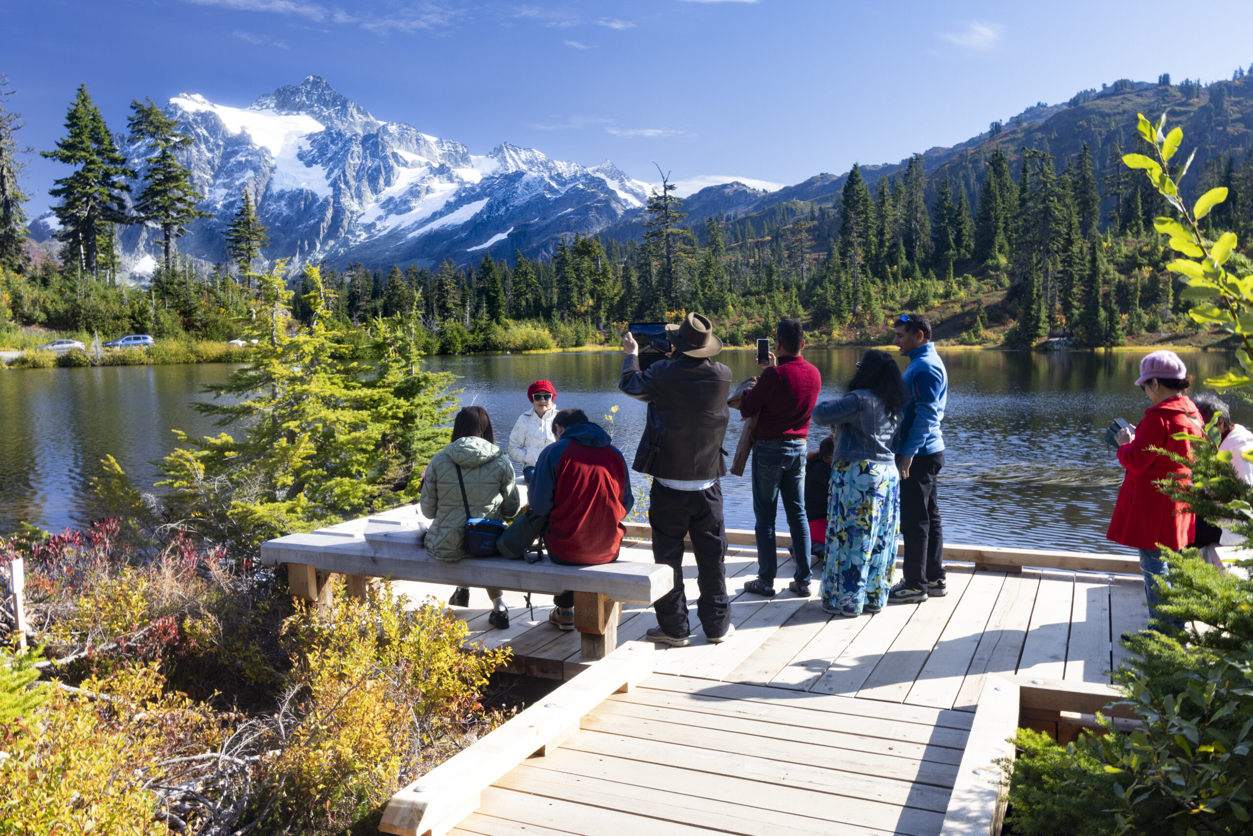 Multiple people visiting a national park. They are standing on a dock next to a body of water and facing a view of a mountain.